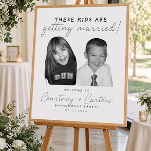 Op de afbeelding: Een rechthoekig wit bord op een houten ezel met de tekst THESE KIDS ARE getting married! boven twee zwart-wit kinderfoto's van een meisje en een jongen. Onder de foto's staat de tekst WELCOME TO Courtney + Carter's ENGAGEMENT PARTY! 03.22.2026.