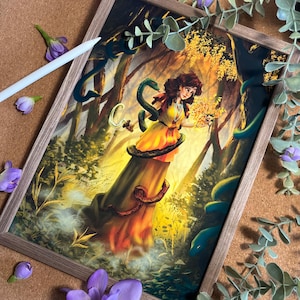 May include: A framed art print featuring a woman in a yellow dress, surrounded by snakes in a forest setting. The artwork has a fantasy theme with a warm color palette, including shades of yellow, orange, and green. The frame is a light brown color.