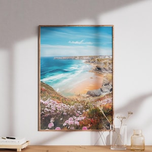 May include: A framed landscape print of a coastal scene. The artwork depicts a beach with turquoise water, a sandy shore, and cliffs. Pink flowers bloom in the foreground. The frame is a warm brown color, and the print is displayed on a wooden cabinet.