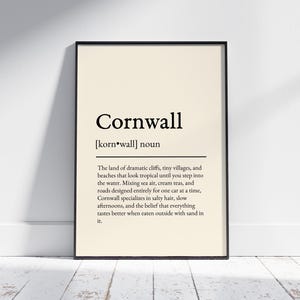 May include: A framed print with the word "Cornwall" in a bold, black font, with a definition below. The background is a light beige color. The frame is black, and the print is leaning against a white wall.