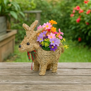 Könnte beinhalten: Ein dekorativer Ziegen-Blumentopf aus Stroh, gefüllt mit bunten Kunstblumen. Die Ziege hat ein rot-grünes Band um den Hals und steht auf einer Holzoberfläche. Die Blumen sind rosa, lila, gelb und orange.