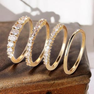 May include: Four gold rings with varying designs. The first ring has oval clear stones, the second has rectangular stones, the third has small round stones, and the fourth is a plain gold band. The rings are displayed on a brown surface.