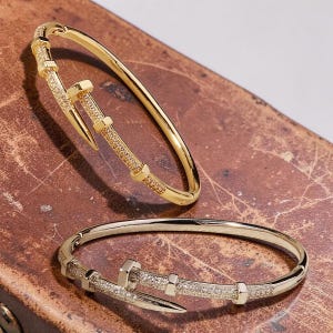 Peut inclure: Deux bracelets joncs articulés, l'un doré et l'autre argenté, arborant un design inspiré d'un clou avec une extrémité pointue et une tête plate, ornés de petites pierres claires le long de l'anneau et dotés d'un fermoir métallique.