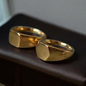 Op de afbeelding: Twee goudkleurige zegelringen. Eén ring heeft een rechthoekige voorkant, de andere een zeshoekige. De ringen zijn gemaakt van glanzend, gepolijst metaal en liggen op een donkerbruin oppervlak. Klassieke sieraden.