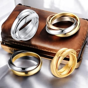Puede incluir: Cuatro anillos tricolores en plata, oro y oro rosa se muestran sobre una superficie de cuero marrón. Los anillos están entrelazados, con tres bandas que forman una sola pieza. Los anillos son una joya.