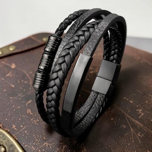 May include: A multi-strand black bracelet featuring a combination of braided leather cords, a smooth flat metal bar, and a cylindrical metal accent piece. The design includes a secure black metal clasp that connects the various textured leather layers.