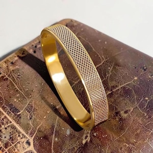 May include: A gold-toned hinged bangle bracelet featuring a wide, flat band with a textured, woven-style mesh pattern on the exterior surface and a smooth, polished interior finish. The bracelet has a secure clasp mechanism visible on the side.