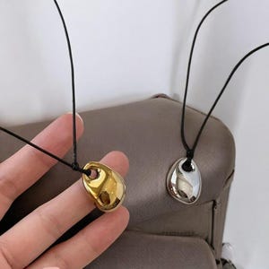 Puede incluir: Dos collares con colgantes de forma orgánica. Un colgante es dorado, el otro plateado. Ambos colgantes cuelgan de un fino cordón negro. Los collares se muestran sobre un fondo neutro.