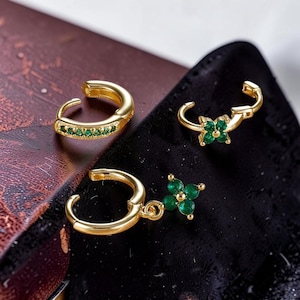 May include: Three gold-colored earrings. One is a hoop with a row of green stones. Another has a flower design with green stones. The third earring has a dangling flower with green stones. The earrings are on a dark background.