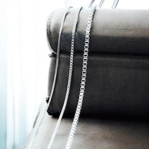 May include: Three silver necklaces with varying chain designs hang against a dark grey backdrop. The chains, composed of small, interconnected links, present a delicate and elegant look. These necklaces are likely jewellery items, suitable for diverse occasions.
