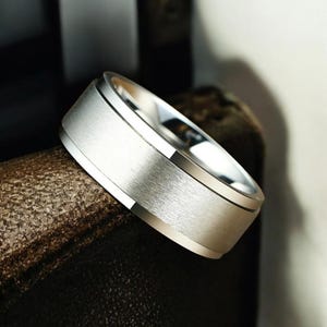 Könnte beinhalten: Ein silberfarbener Ring mit gebürsteter Metalloberfläche. Der Ring hat ein breites Band mit polierter Kante und strukturierter Mitte. Der Ring wird auf einer braunen Oberfläche präsentiert.