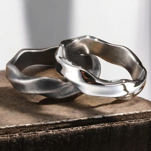 May include: Two silver rings with a wavy, organic design. One ring is a darker, matte silver, while the other is polished and reflective. The rings are interlocked, showcasing their unique, textured surfaces. The rings are displayed on a brown surface.