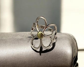 May include: A silver-colored ring with a flower design. The ring features five curved petals and a center with a textured, gold-colored appearance. The ring is displayed on a gray surface.