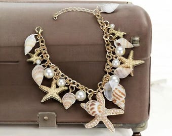 Op de afbeelding: Een goudkleurige bedelarmband met zeeschelpen, zeesterren en imitatieparels. De armband heeft meerdere strengen en is versierd met verschillende maten en vormen van zeeschelpen en zeesterren. De armband wordt gepresenteerd op een bruin oppervlak.