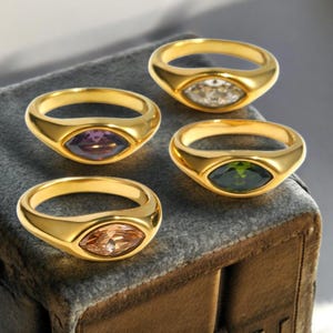 May include: Four gold-colored rings with oval-shaped gemstones in various colors: clear, purple, green, and peach. The rings are displayed on a gray, textured surface, showcasing their polished finish and gemstone details. The rings are a fashion accessory.