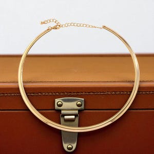 May include: A gold choker necklace with a minimalist design. The necklace is a rigid, circular band that sits close to the neck, with an adjustable chain clasp. The jewelry is displayed on a brown leather surface.