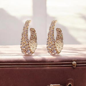 May include: A pair of gold-toned hoop earrings encrusted with sparkling crystals. The earrings are crescent-shaped and appear to be made of metal. The earrings are displayed on a brown surface.