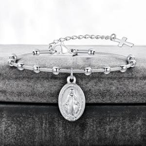 Puede incluir: Una pulsera plateada con pequeñas cuentas redondas y un colgante ovalado que representa una figura religiosa. Un pequeño dije de cruz está unido a la cadena ajustable. La pulsera se exhibe sobre una superficie gris.