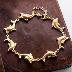 May include: A gold-colored bracelet featuring dolphin charms. The bracelet has a chain-link design with seven dolphin pendants. Each dolphin has a small, round accent. The bracelet is displayed on a brown surface.