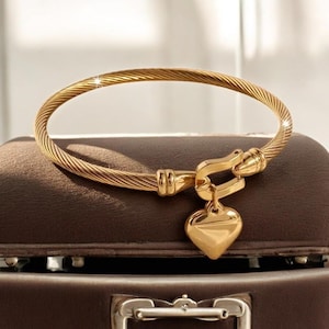Puede incluir: Un brazalete dorado con un diseño de cuerda trenzada. La pulsera presenta un dije en forma de corazón y un cierre de broche. La joya se exhibe sobre una superficie marrón.