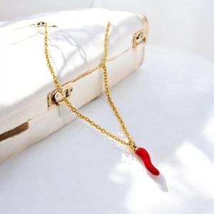 May include: A gold chain necklace featuring a small, red chilli pepper pendant. The necklace is set against a white surface and a white chest. The pendant is a bright red colour.