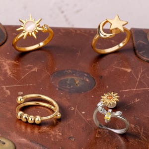 Könnte beinhalten: Vier Ringe mit von Himmel und Natur inspirierten Designs. Ein Ring zeigt eine Sonne mit Perlenmitte, ein anderer einen Mond und Stern, ein dritter ein Doppelband mit kleinen Kugeln und der letzte eine Sonnenblume und eine Libelle.