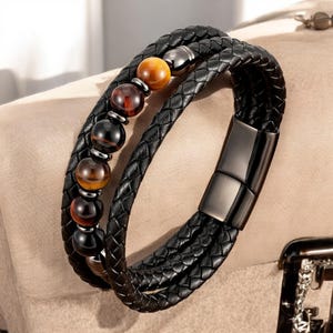 May include: Black braided leather bracelet with a magnetic clasp. The bracelet features a row of polished, round beads in shades of brown, amber, and black. The bracelet is designed with multiple strands of braided leather.