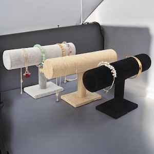 May include: Four jewelry display stands in gray, beige, and black. Each stand has a horizontal, cylindrical bar for displaying bracelets and necklaces. The stands are made of different materials, including velvet and linen, and are holding various bracelets.