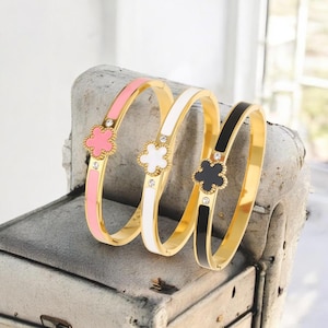 Può includere: Tre braccialetti color oro con dettagli a forma di quadrifoglio. Un braccialetto è rosa, uno bianco e uno nero. Ogni braccialetto presenta un quadrifoglio con un piccolo cristallo al centro. I braccialetti sono esposti su una superficie strutturata.