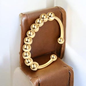 Puede incluir: Un anillo dorado con una serie de pequeñas cuentas redondas conectadas en una fila. El anillo tiene un diseño curvo con una cuenta en cada extremo. El anillo se muestra sobre una superficie de cuero marrón.