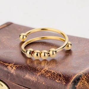 May include: A gold-colored ring with a spiral design. The ring features four small, round, gold-colored beads that can be moved along the band. The ring is shown on a brown, textured surface.