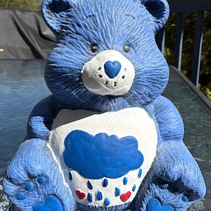 May include: A blue bear figurine with a white belly featuring a blue cloud with raindrops and red hearts. The bear has a heart-shaped nose and paw pads. The figurine is painted in shades of blue and white.