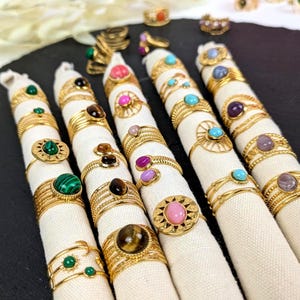 Op de afbeelding: Een verzameling goudkleurige ringen met diverse edelstenen, waaronder groene malachiet, zwarte onyx, roze en turkoois stenen. De ringen worden gepresenteerd op witte stoffen rollen tegen een donkere achtergrond. De ringen hebben ingewikkelde ontwerpen.