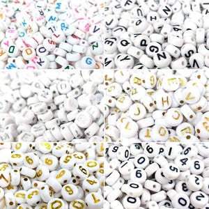 Könnte beinhalten: Eine Sammlung weißer, runder Perlen mit aufgedruckten Buchstaben und Zahlen in verschiedenen Farben und Schriftarten. Einige Perlen haben schwarze Buchstaben, andere goldene Zahlen und Buchstaben. Für Bastelarbeiten und Schmuckherstellung.