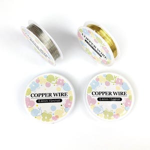 May include: Four spools of copper wire are displayed against a white background. Two spools are silver, and two are gold. The spools are labeled "COPPER WIRE" and "0.4mm 15m/roll" with a floral design.