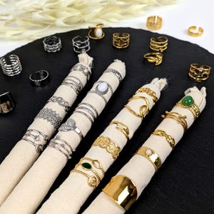 Op de afbeelding: Een assortiment ringen in zilver- en goudtinten, gepresenteerd op crèmekleurige stoffen rollen tegen een zwarte achtergrond. De ringen hebben verschillende ontwerpen, waaronder banden, textuurpatronen en edelsteenaccenten. Sommige ringen zijn gestapeld, wat een verscheidenheid aan stijlen laat zien.