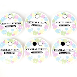 Könnte beinhalten: Sechs runde Etiketten mit der Aufschrift "CRYSTAL STRING" und Maßen. Jedes Etikett zeigt ein Blumen- und Schneckenmuster in Pastellfarben. Die Etiketten haben unterschiedliche Fadendicken, von 0,4 mm bis 1,0 mm, und Längen von 10 m bis 20 m.