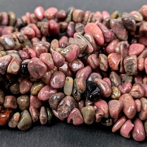 Könnte beinhalten: Nahaufnahme einer Sammlung getrommelter Edelsteine in Rosa-, Braun- und Grautönen. Die Steine variieren in Größe und Form und haben eine polierte, leicht glänzende Oberfläche. Die Edelsteine sind aufgefädelt, was darauf hindeutet, dass sie für die Schmuckherstellung bestimmt sind.