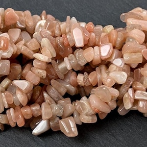 Könnte beinhalten: Nahaufnahme eines Strangs aus Sonnenstein-Chip-Perlen. Die Perlen sind eine Mischung aus Pfirsich-, Beige- und Weißtönen. Die Chips sind unregelmäßig geformt und unterschiedlich groß, wodurch ein strukturiertes Aussehen entsteht. Ideal für die Schmuckherstellung.