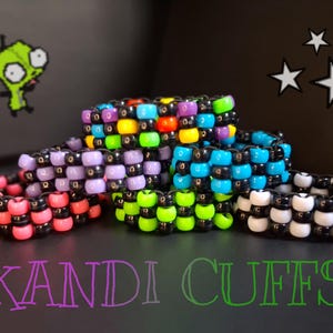 Può includere: Una collezione di colorati braccialetti Kandi, impilati e disposti su una superficie scura. I braccialetti presentano varie combinazioni di colori, tra cui perline rosa, nere, blu, verdi e viola. Il testo "KANDI CUFFS" è visualizzato in gradiente.
