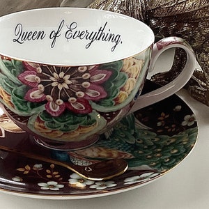 May include: A white teacup and saucer with a floral design. The teacup has the words "Queen of Everything" written on the inside. The saucer has a peacock design.