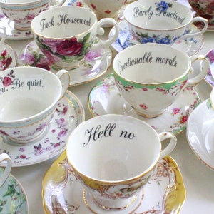 May include: A collection of teacups with humorous and sarcastic phrases printed on them. The teacups are white with floral patterns and gold trim. The phrases include "Shitbird", "Fuck Housewerk", "Barely Functioned", "Drama Queen", "Questionable morals", "I love you. Be quiet", "Hell no", and "Dr. Stupid".