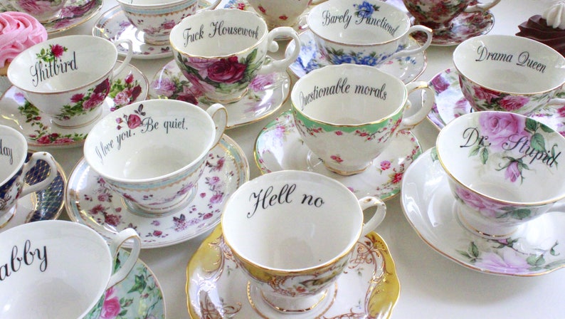 May include: A collection of teacups with humorous and sarcastic phrases printed on them. The teacups are white with floral patterns and gold trim. The phrases include "Shitbird", "Fuck Housewerk", "Barely Functioned", "Drama Queen", "Questionable morals", "I love you. Be quiet", "Hell no", and "Dr. Stupid".