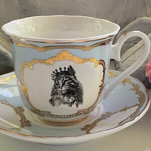 K&ouml;nnte beinhalten: Ein hellblaues Tee-Set mit goldenen Verzierungen. Die Teetasse zeigt eine Schwarzwei&szlig;-Illustration einer Katze mit einer Krone. Die Untertasse hat ein &auml;hnliches Design mit goldenen Akzenten.