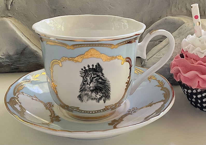 K&ouml;nnte beinhalten: Ein hellblaues Tee-Set mit goldenen Verzierungen. Die Teetasse zeigt eine Schwarzwei&szlig;-Illustration einer Katze mit einer Krone. Die Untertasse hat ein &auml;hnliches Design mit goldenen Akzenten.