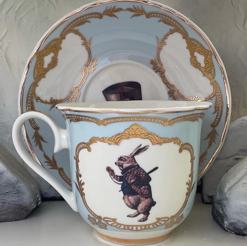 Puede incluir: Un juego de taza y platillo de t&eacute; azul claro con ribetes dorados. La taza de t&eacute; presenta una ilustraci&oacute;n de un conejo blanco con chaleco y reloj de bolsillo. El platillo tiene un dise&ntilde;o dorado con una peque&ntilde;a imagen de un sombrero de copa.