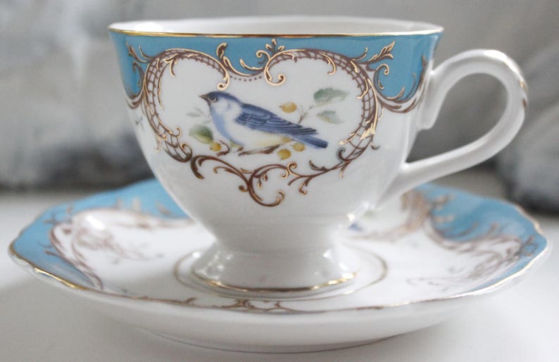 May include: A blue and white teacup and saucer set with a gold trim and a bluebird design.