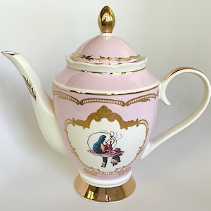 Puede incluir: Una tetera de porcelana rosa con detalles dorados y una ilustraci&oacute;n caprichosa de una mujer sentada en un hongo. La tetera tiene un pico y un asa blancos.
