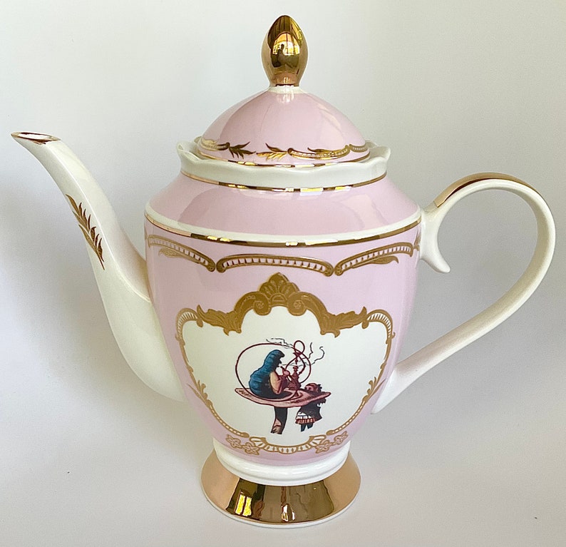 Puede incluir: Una tetera de porcelana rosa con detalles dorados y una ilustraci&oacute;n caprichosa de una mujer sentada en un hongo. La tetera tiene un pico y un asa blancos.