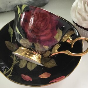 Puede incluir: Un juego de taza y platillo de t&eacute; negro con borde dorado. La taza y el platillo presentan una gran rosa roja con hojas verdes y detalles dorados.